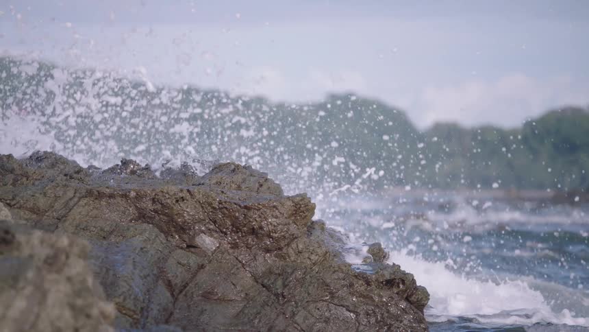 A powerful ocean wave crashes against a jagged coastal rock formation in dramatic slow motion