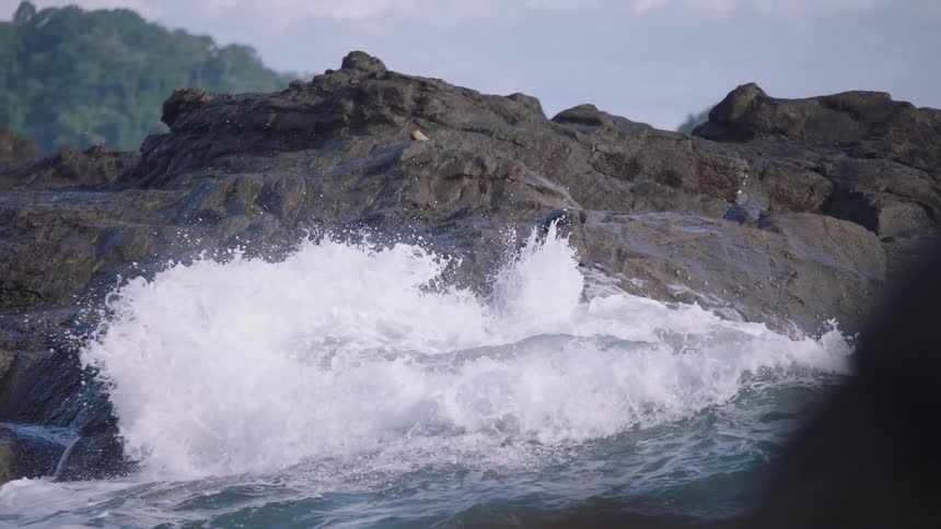 Powerful ocean waves crash against dark, wet rocks on a tropical shoreline in slow motion