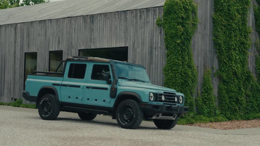 A modern teal off-road truck parks on a gravel driveway in front of a rustic wooden building covered in ivy