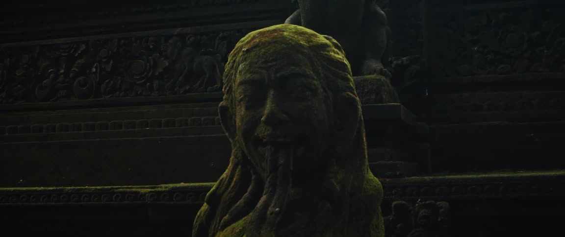 A slow zoom out reveals an ancient, moss-covered stone statue of a demonic figure at a dark, mystical temple in Bali