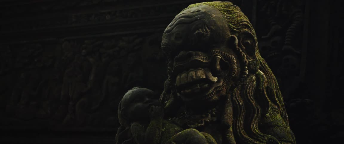 A slow zoom out on an ancient, moss-covered stone statue of a mythological creature holding a baby at a temple in Bali