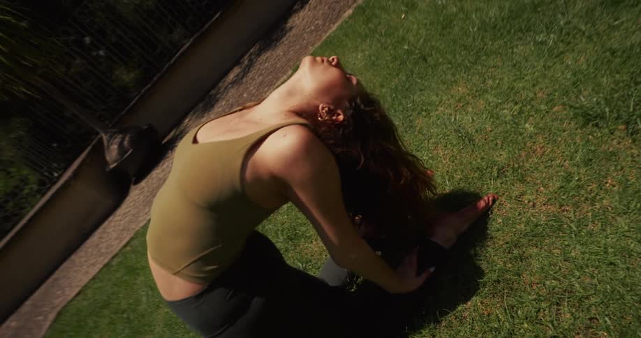 A high-angle view shows a woman in a deep backbend yoga pose, stretching on a sunny green lawn outdoors