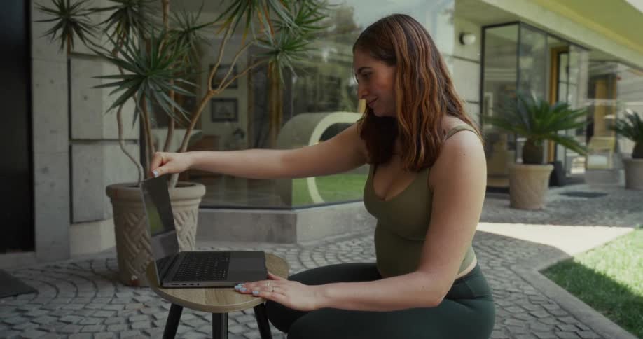 A woman in athletic wear opens a laptop on the patio of her home in slow motion