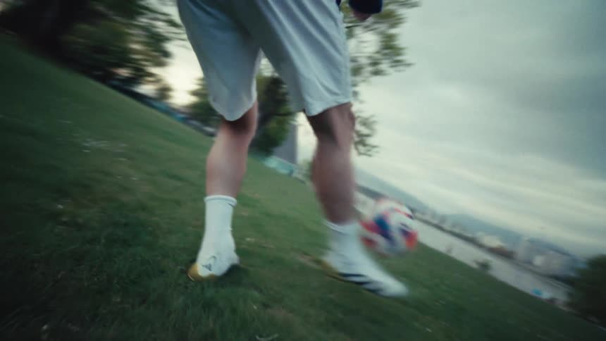 A soccer player skillfully dribbles and juggles a ball down a grassy hill in a park, captured in an immersive slow-motion, low-angle shot