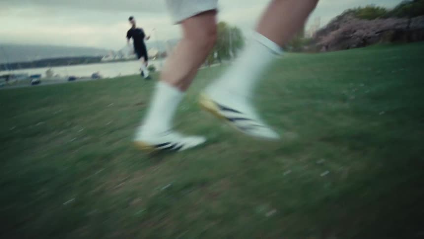 A low-angle, slow-motion shot follows a soccer player's feet as they run and dribble a ball across a grassy park