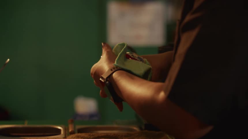 A barista meticulously wipes down a hand-painted ceramic face mug with a green cloth in a dimly lit cafe