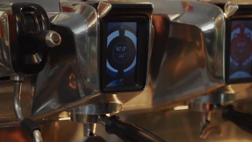 A barista's hand with black nail polish and a crescent moon tattoo programs a modern chrome espresso machine using its digital touchscreen