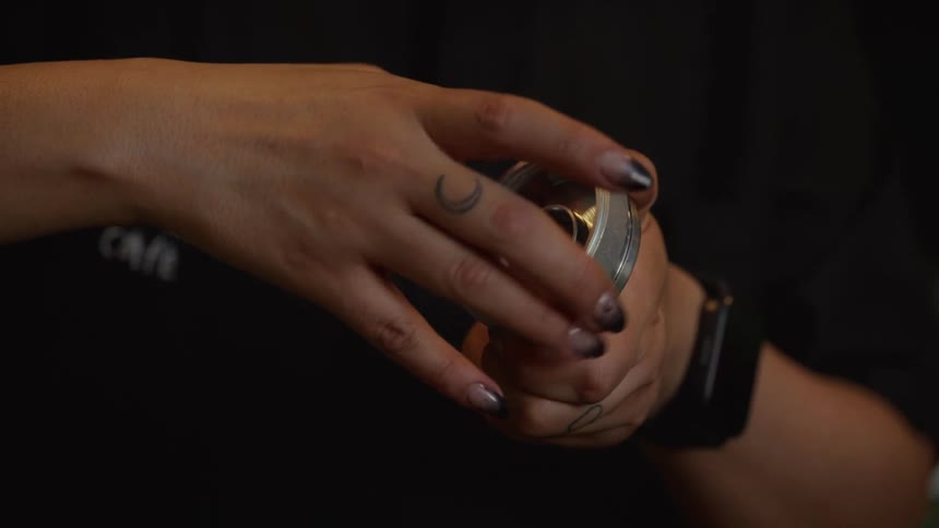 A barista's tattooed hands slowly turn a manual coffee grinder, preparing fresh grounds