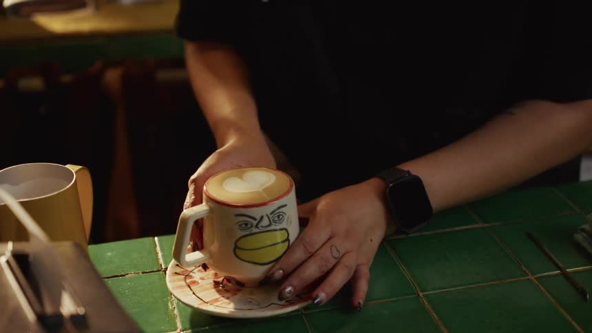 A barista carefully places a freshly made latte with heart art in a unique hand-painted mug on a green tiled cafe counter
