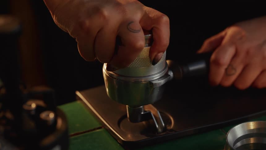 Close-up slow-motion shot of a barista with tattoos tamping fresh ground coffee in a portafilter