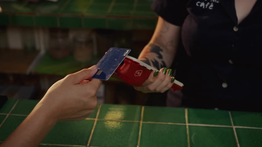 A customer makes a payment by slowly sliding a credit card into a portable terminal held by a cafe employee