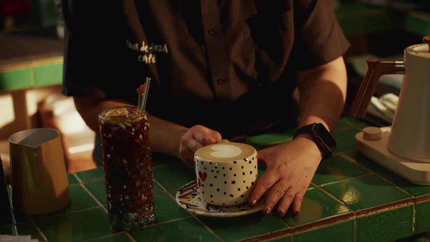A barista carefully presents a freshly made latte with heart-shaped art in a polka-dotted mug at a cozy cafe