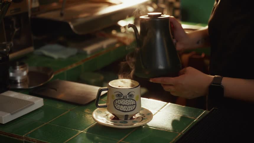 A barista pours steaming hot water into a quirky, cartoon-faced mug on a green-tiled counter, all captured in beautiful slow motion