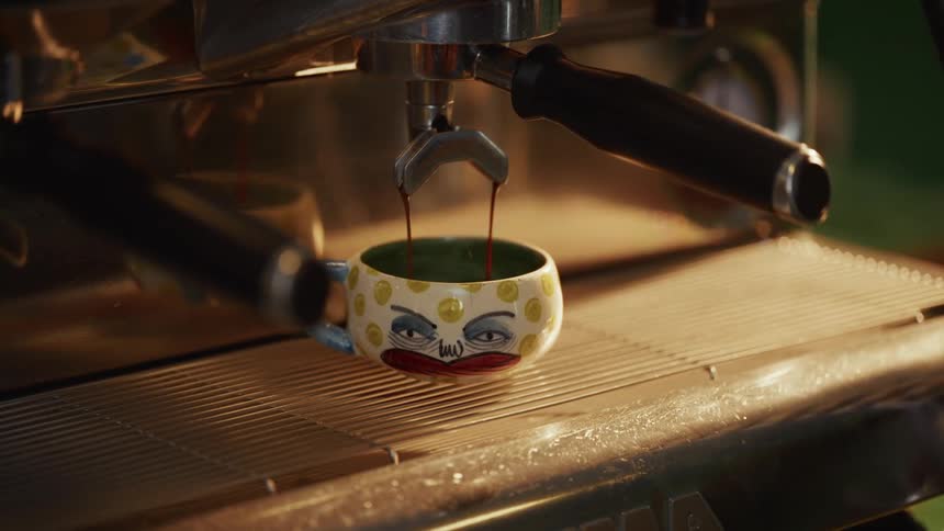 A close-up shot of a chrome espresso machine pouring fresh hot coffee into a quirky, artistic, hand-painted mug