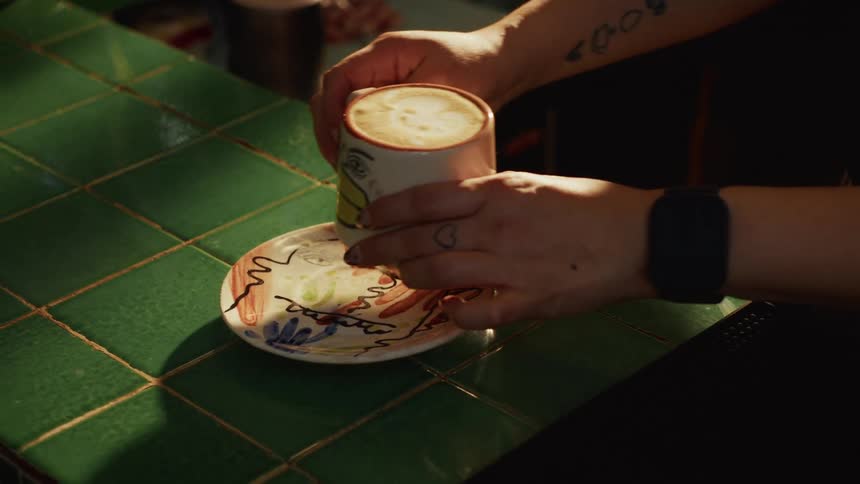A tattooed barista carefully places a cappuccino with latte art in a quirky, artistic mug onto a matching saucer on a green tiled counter