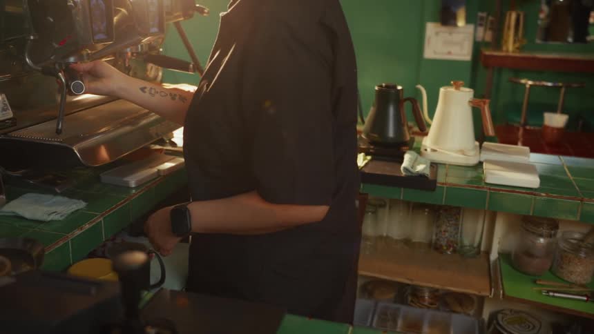 A barista with a tattoo on their arm operates an espresso machine behind a green tiled counter in a sunlit cafe