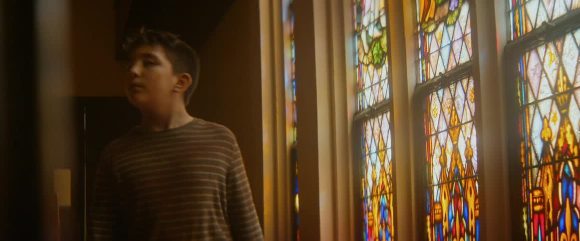 A curious young boy walks into a sunlit cathedral and gazes up in wonder at the vibrant stained-glass windows