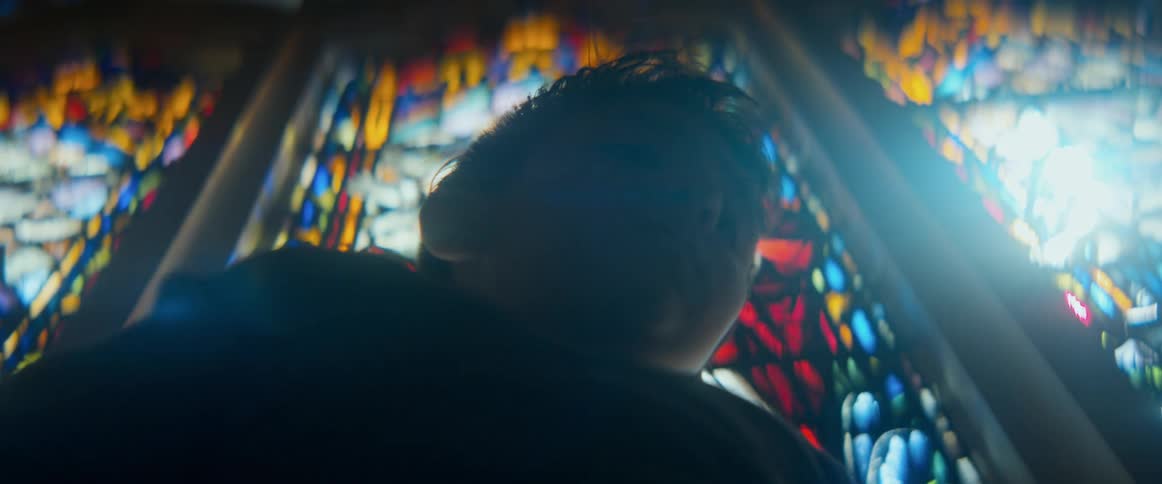 A low-angle shot captures a silhouetted figure bowing their head in prayer before a vibrant stained-glass window in a church