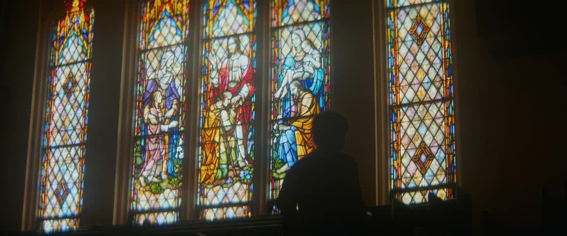 A silhouetted person sits in a church pew, contemplating a vibrant stained-glass window depicting biblical scenes