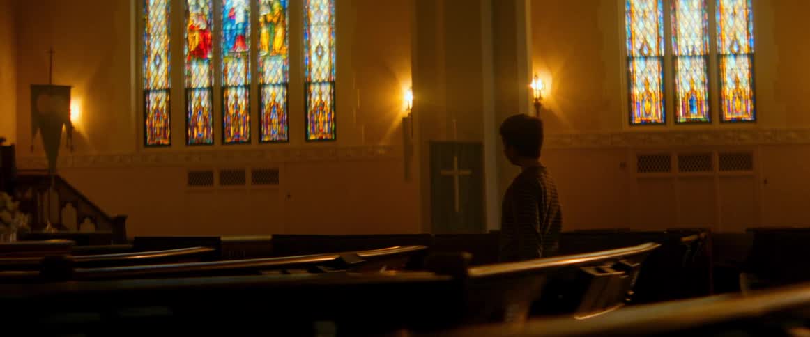 A young boy stands alone in a quiet church, gazing up at the beautiful stained glass windows