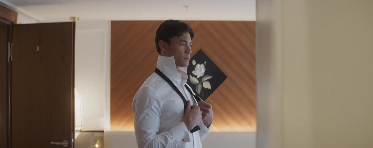 A handsome man in a white dress shirt carefully adjusts his black bow tie while preparing for a formal event in a stylish room