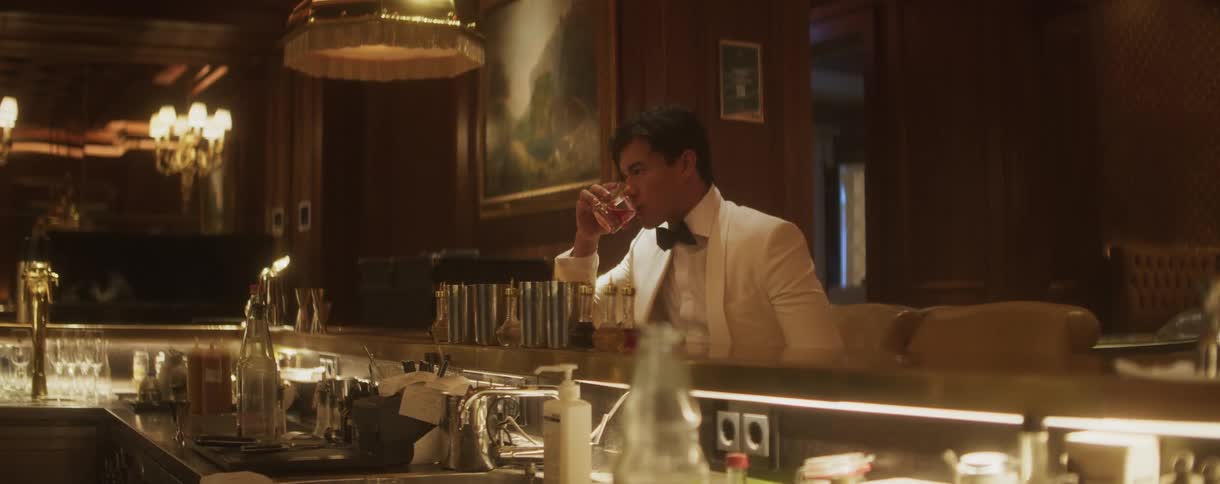 A sophisticated gentleman in a white tuxedo jacket enjoys a cocktail alone at an elegant, dimly lit bar
