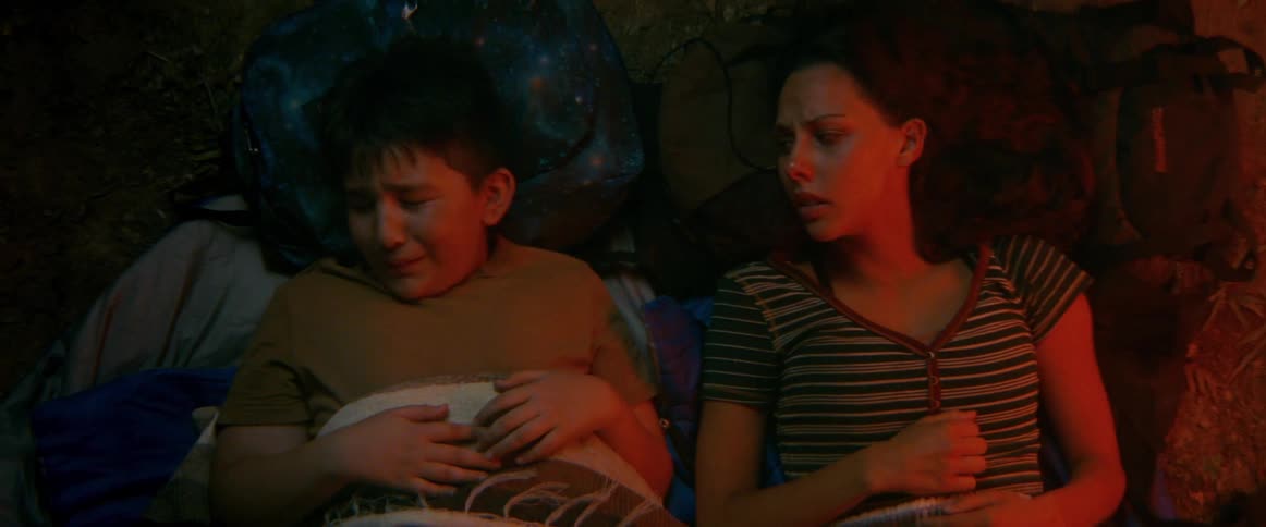Worried teenage girl comforts a crying boy while lying under blankets in a dim red-lit campsite