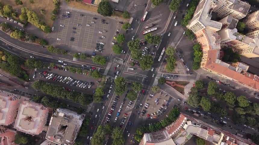 A stunning top-down aerial shot reveals a bustling city intersection, with cars and buses navigating through streets lined with trees and historic buildings as the camera slowly zooms out