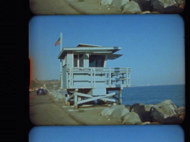 An 8mm film scan of an empty lifeguard tower with an American flag standing by the ocean on a sunny day