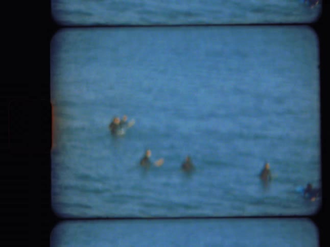 A vintage film captures blurry surfers floating in the blue ocean and a close-up of a seagull on the rocks