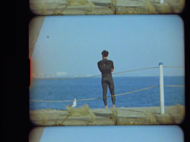 Vintage Super 8 footage shows a man in a wetsuit standing on a pier, looking out at the vast blue ocean on a sunny day