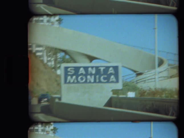 A vintage Super 8mm film scan shows the iconic Santa Monica sign along a sunny California highway
