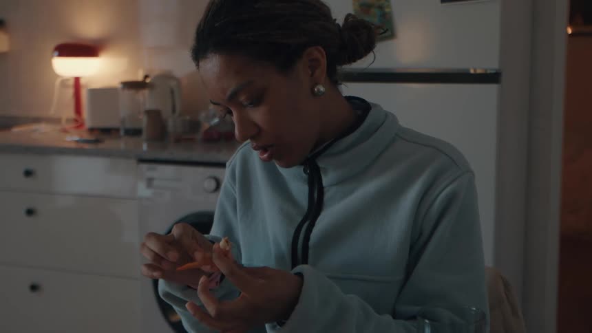 A young woman with a focused expression carefully peels a fresh shrimp in her kitchen at night