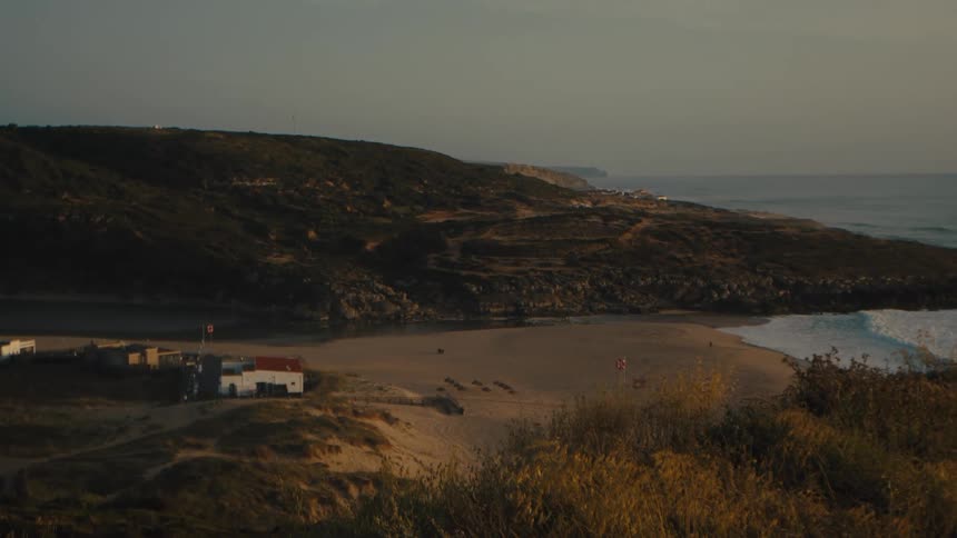 A secluded sandy beach cove with a few small buildings is bathed in the warm light of golden hour
