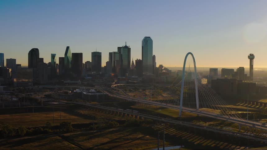 A serene aerial view shows the Dallas skyline and the Margaret Hunt Hill Bridge bathed in the warm golden light of sunrise