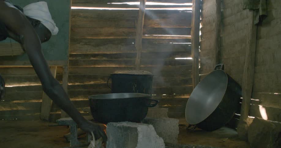 A person tends to a cooking fire in a simple wooden shack as sunlight streams through the walls