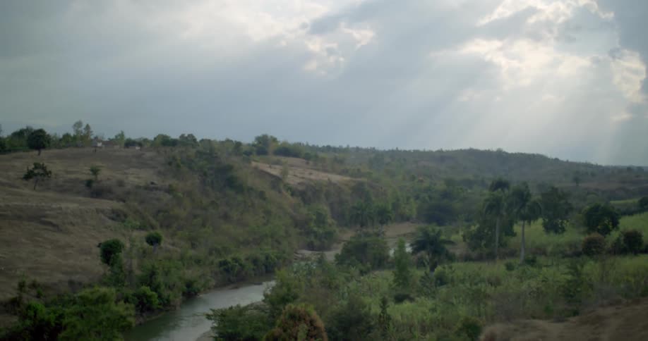 A slow-moving shot reveals a winding river through a lush, hilly landscape as sunbeams break through the clouds