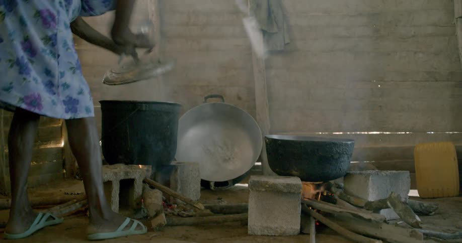 A woman cooks food in large pots over an open fire inside a rustic wooden kitchen in a developing country