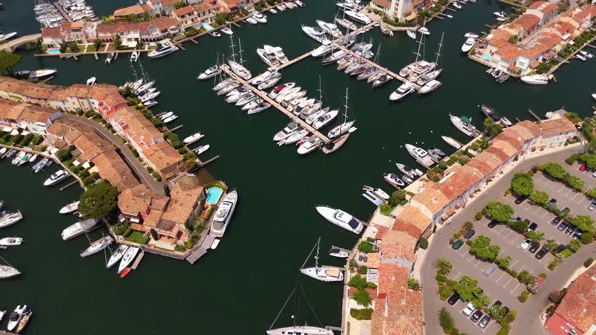 An aerial drone shot slowly ascends over a picturesque and luxurious European marina filled with docked sailboats and yachts on a sunny day