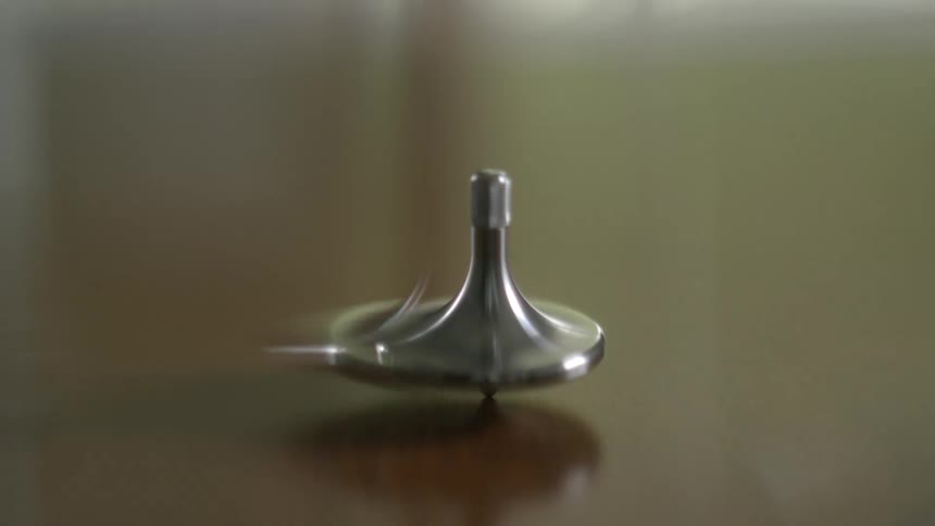 A sleek metallic spinning top rotates with motion blur before slowing to a wobble on a smooth, reflective table
