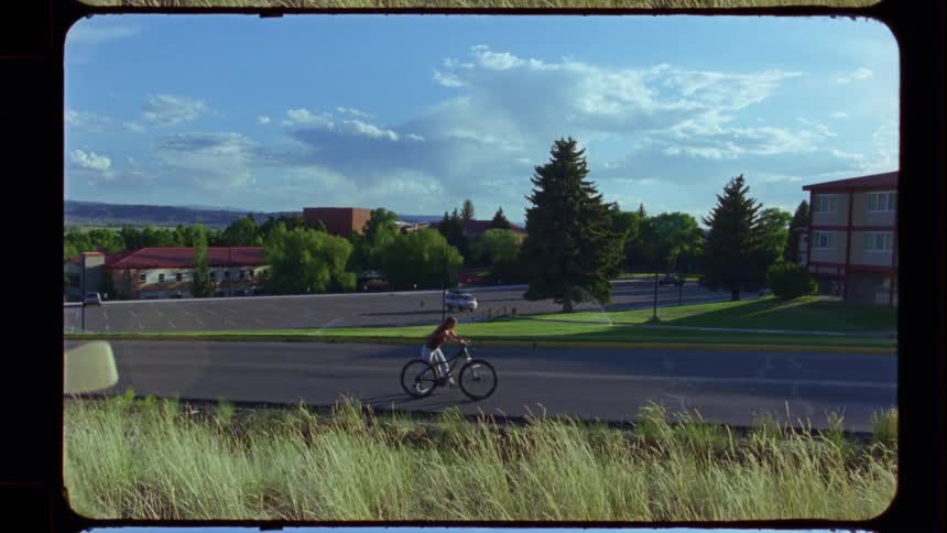 A person on a bicycle rides down a road in a quiet suburban town on a sunny day, captured on vintage film