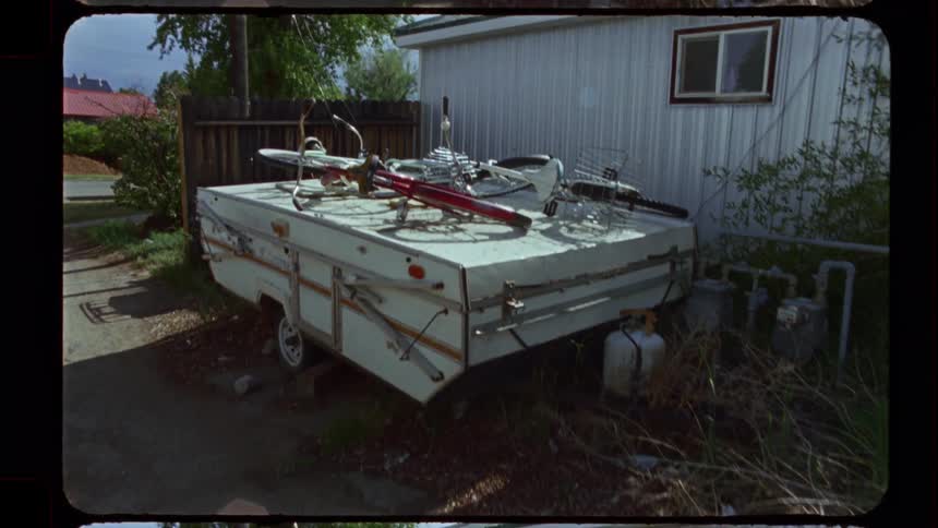 An old, forgotten pop-up camper trailer sits in a weedy backyard with a disassembled bicycle on its roof