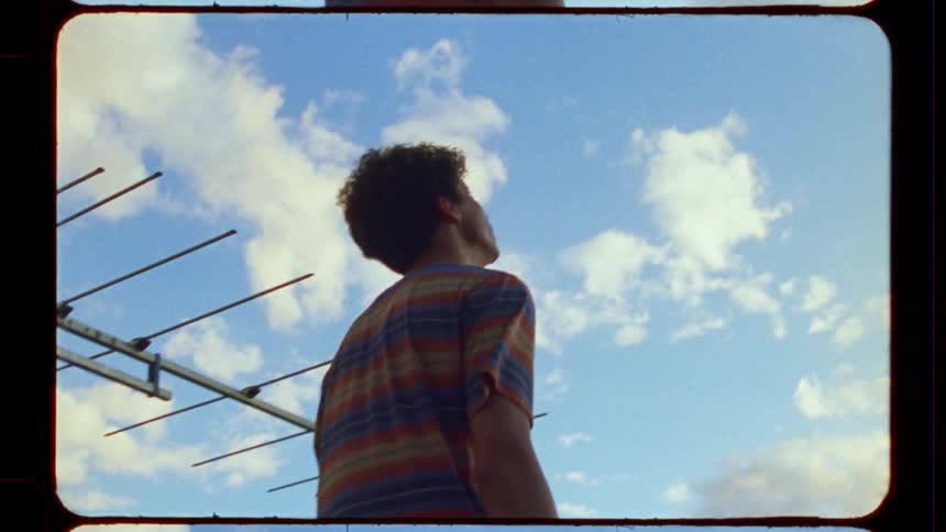 A dreamy young man gazes thoughtfully at the cloudy blue sky from a rooftop, with a television antenna in the foreground