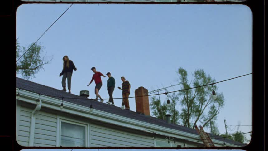 Vintage film shows a group of adventurous young men carefully walking in a line along the peak of a house roof