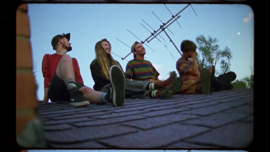 A group of four young friends laugh and talk while relaxing on a suburban rooftop at dusk in this nostalgic, vintage-style film clip