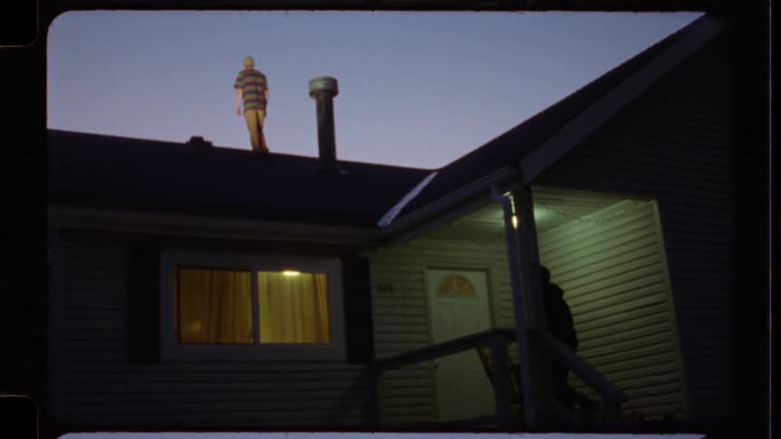 A man stands on a house roof at dusk as a silhouetted figure watches from the porch below in a surreal, vintage film-style shot