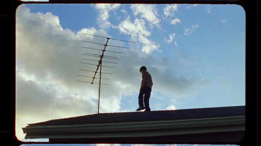 A young boy in silhouette walks across a rooftop and stands still, framed by a dramatic cloudy sky on vintage film