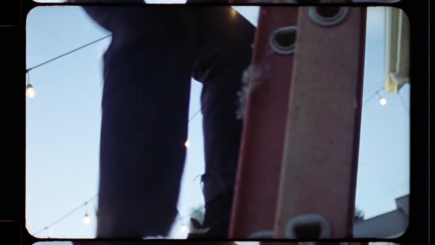 A young man in a striped t-shirt carefully climbs down a red ladder in his backyard, captured on vintage film