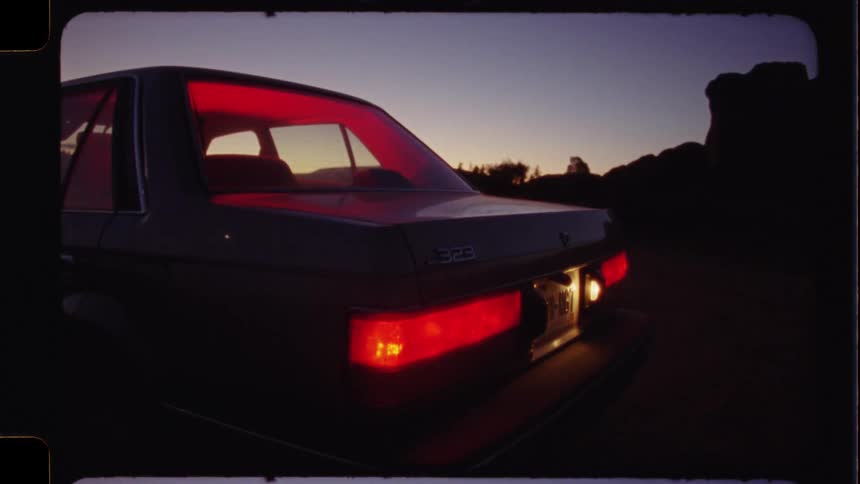 The rear lights of a vintage sedan glow red as day breaks in a desolate desert landscape, captured on vintage film
