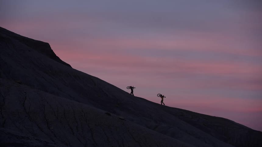 Two silhouetted mountain bikers carry their bikes up a steep, dark mountain against a vibrant pink and purple sunset sky